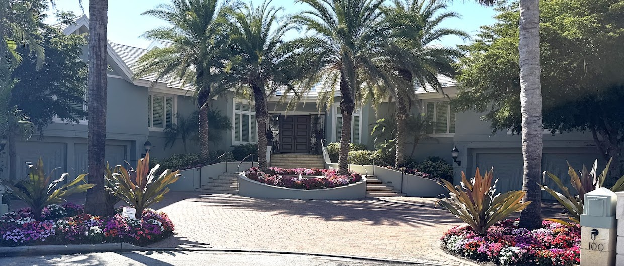 Coastal estate entrance framed by mature palms, with clipped hedging, seasonal flower beds and a paver motor court.